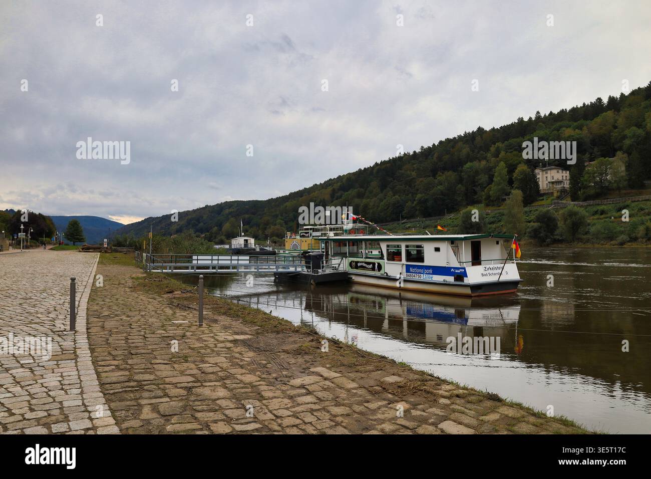Personenfähre a Bad Schandau am Anleger: Blick über die gepflasterte Uferpromenade auf das Fährschiff vor bewaldeten Elbhängen a Sachsen Foto Stock