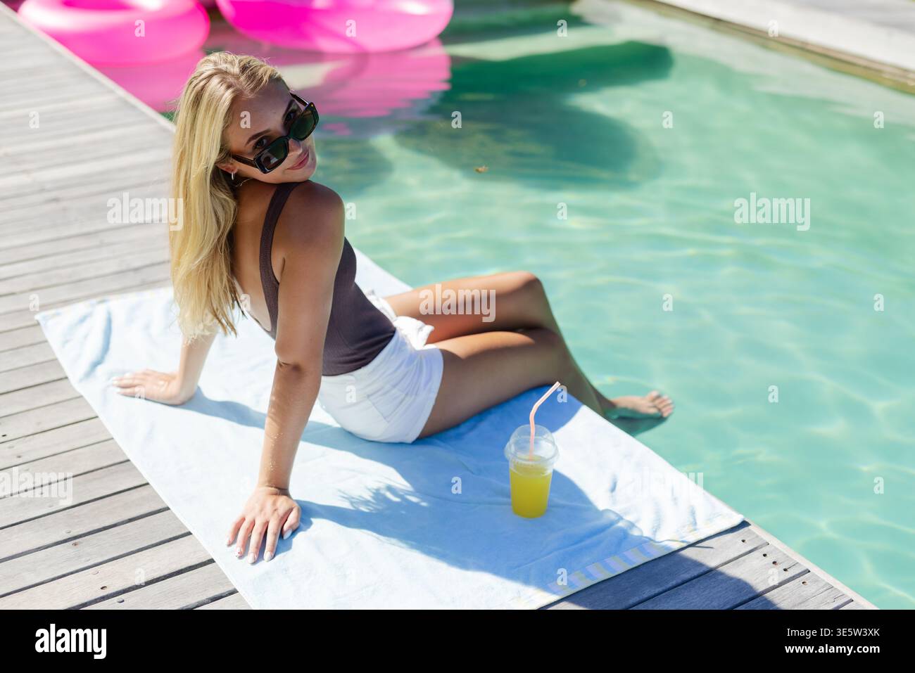 Donna adulta seduta su un asciugamano sulla terrazza in legno della piscina, piede subacqueo, costume da bagno, occhiali da sole, tazza trasparente Foto Stock