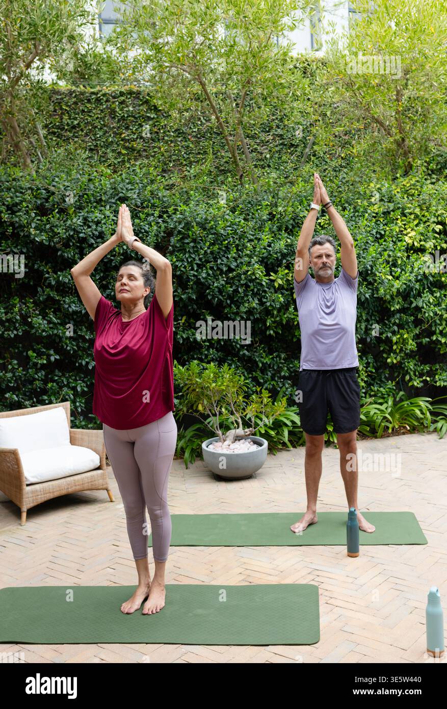 Coppie anziane diverse che fanno yoga in piedi su tappeti verdi sul patio indossando un top Bordeaux con bottiglie Foto Stock