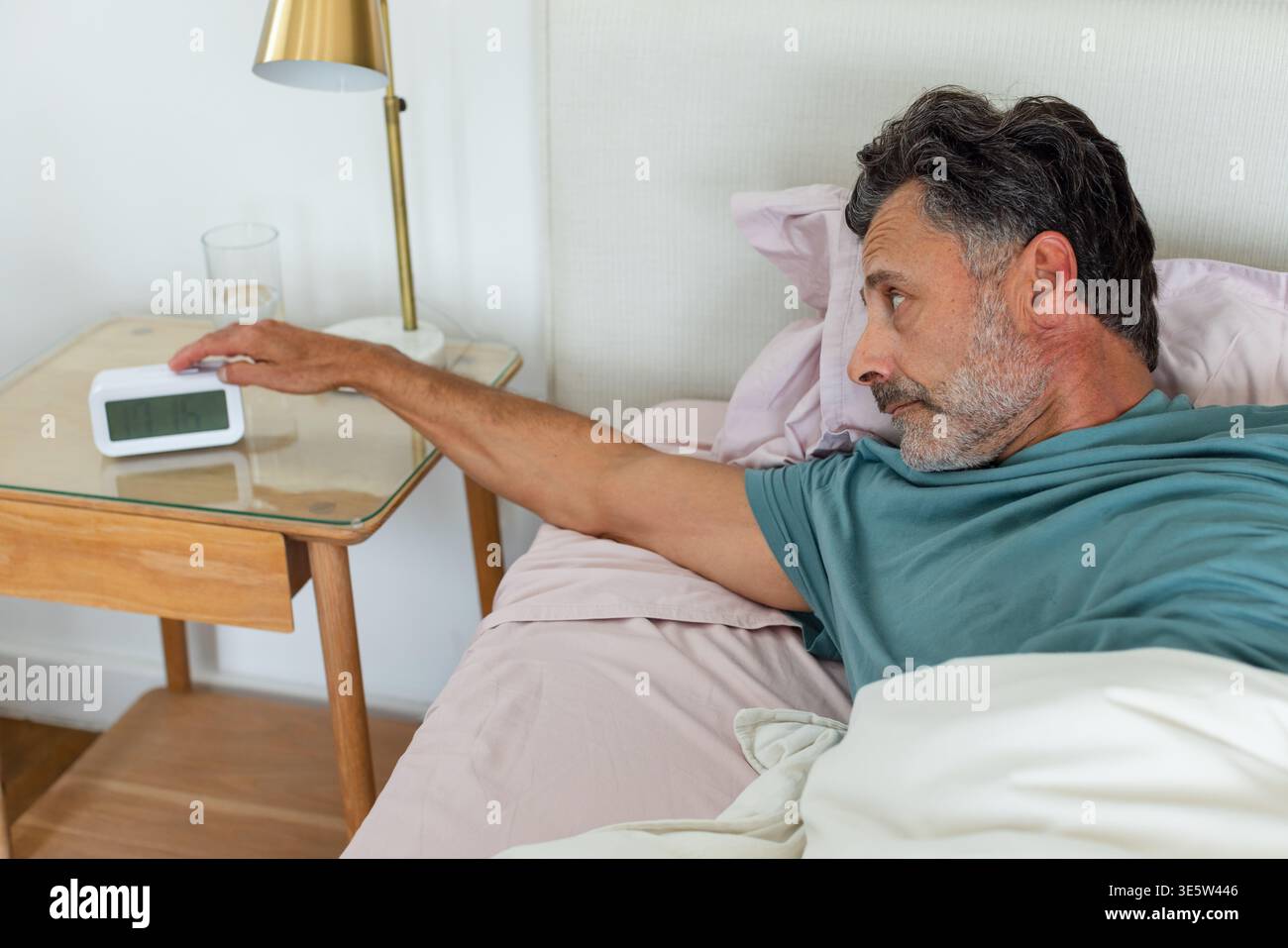 Uomo di mezza età sdraiato su lenzuola rosa in camera da letto che arriva verso la sveglia digitale con una camicia color ottanio Foto Stock