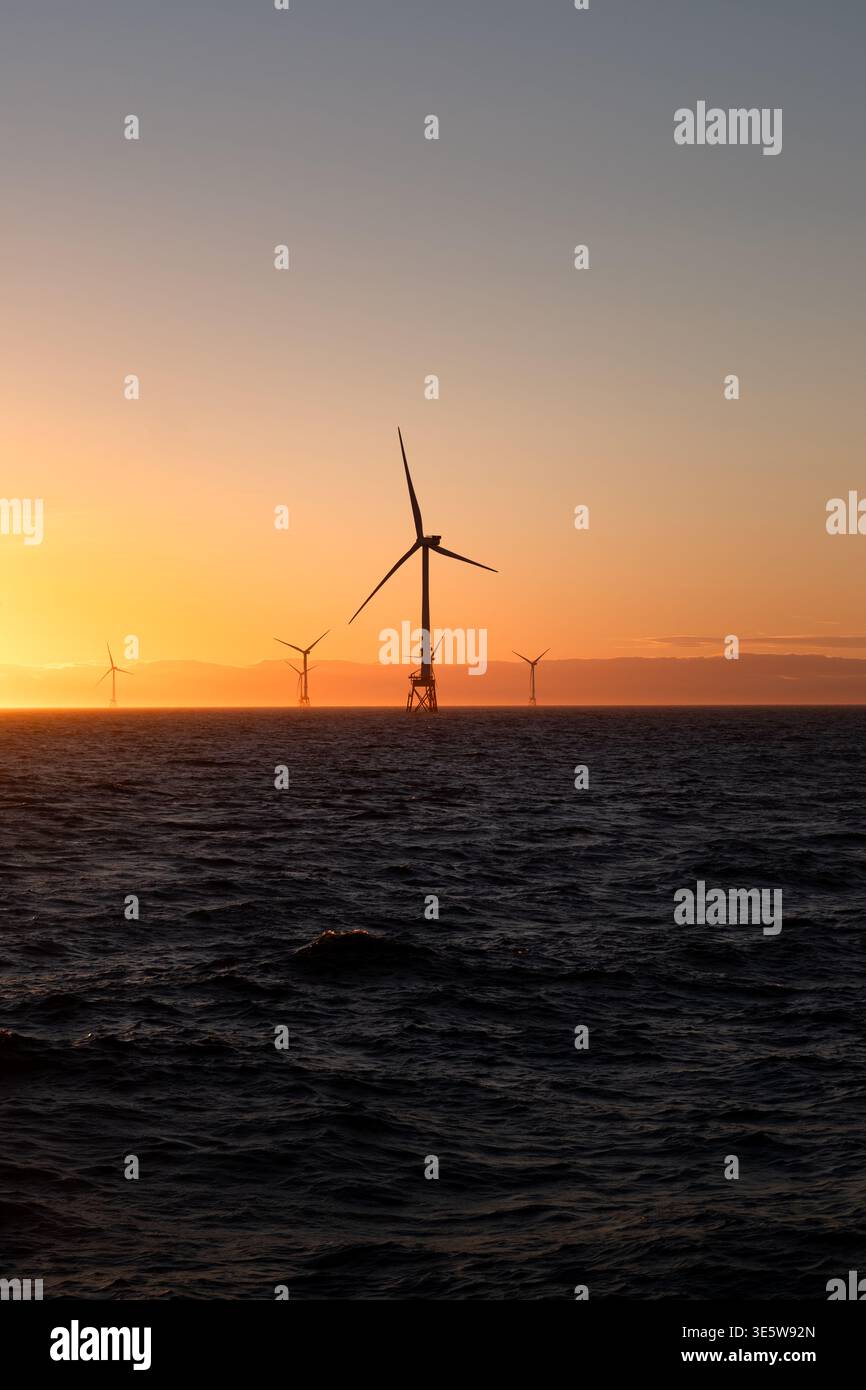 Una fila di turbine eoliche offshore si staglia contro un vivace tramonto arancione e dorato sul Mare del Nord, con colline costiere visibili oltre. Foto Stock
