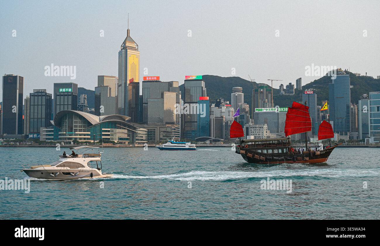 Tradizionale junk boat cinese con vele rosse che navigano attraverso Victoria Harbour con lo skyline di Hong Kong sullo sfondo. Foto Stock