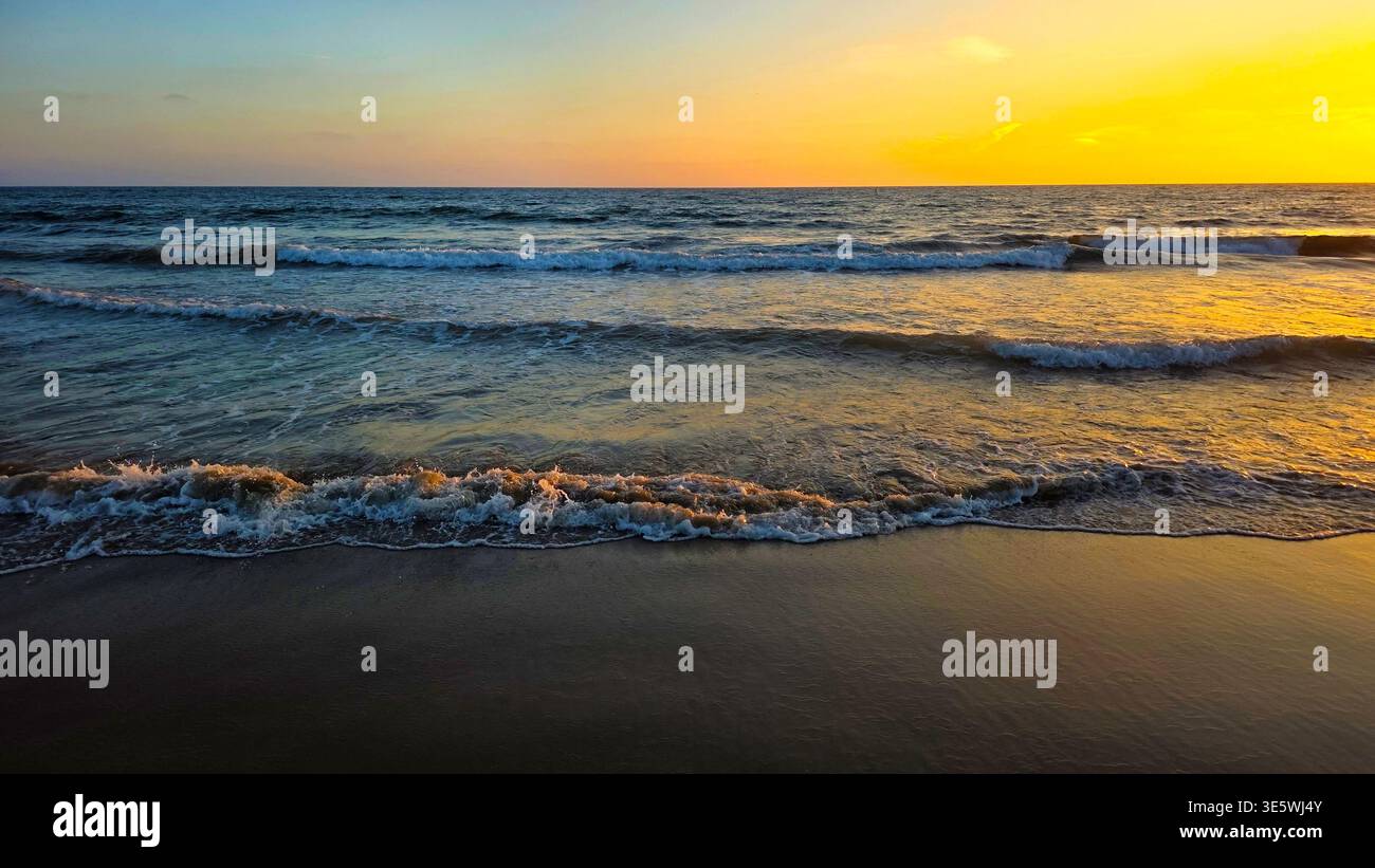 Il Golden Sunset Waves si snoda su Sandy Beach. La luce dorata del tramonto si riflette sulle onde dolci e sulla sabbia bagnata, creando un tranquillo paesaggio marino lungo la costa Foto Stock