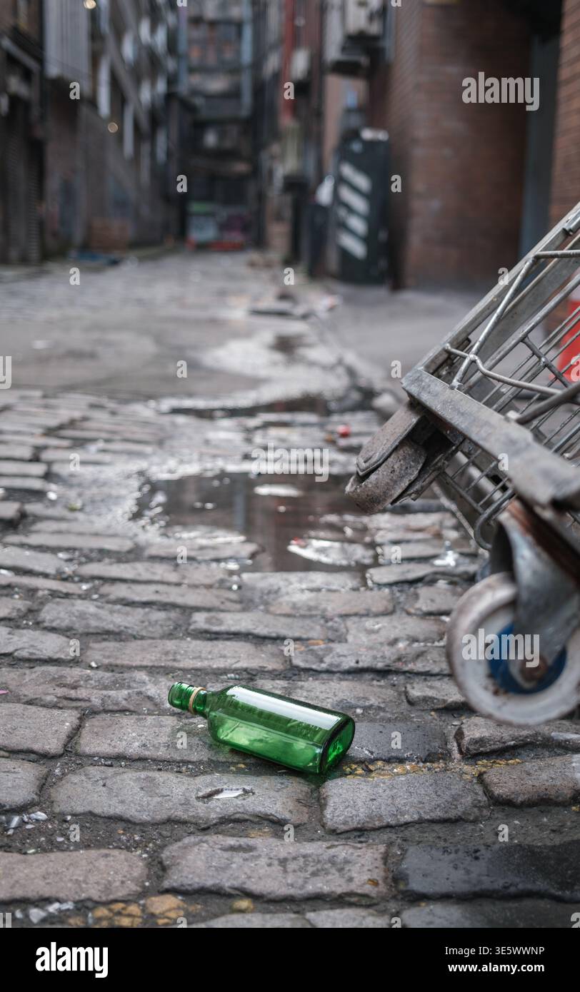 Un tram rovesciato e una bottiglia di vetro verde gettata su ciottoli umidi In Uno stretto vicolo urbano a Glasgow Foto Stock