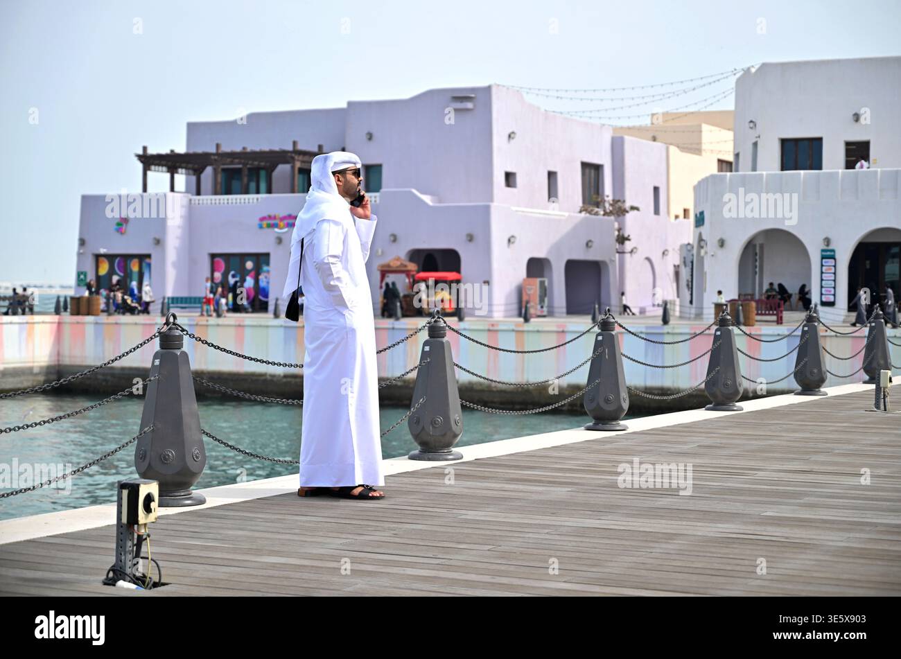 Qatar, Doha : uomo del Qatar che indossa abiti tradizionali, sandali e occhiali da sole, parla al cellulare su un molo nel distretto di Mina Foto Stock