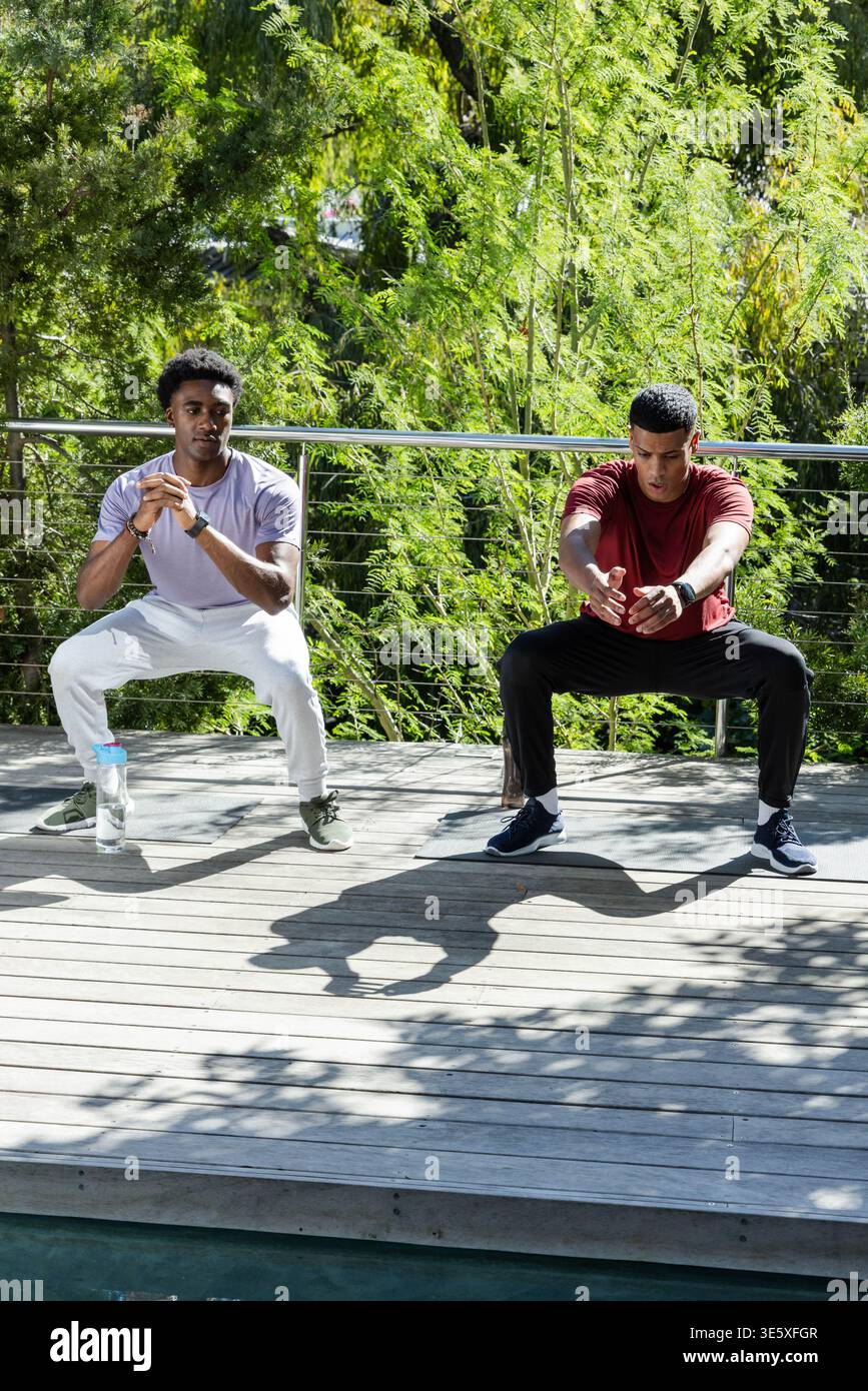 Accovacciare diversi amici maschi che eseguono la routine di fitness sul ponte, con una bottiglia d'acqua e dei tracker Foto Stock