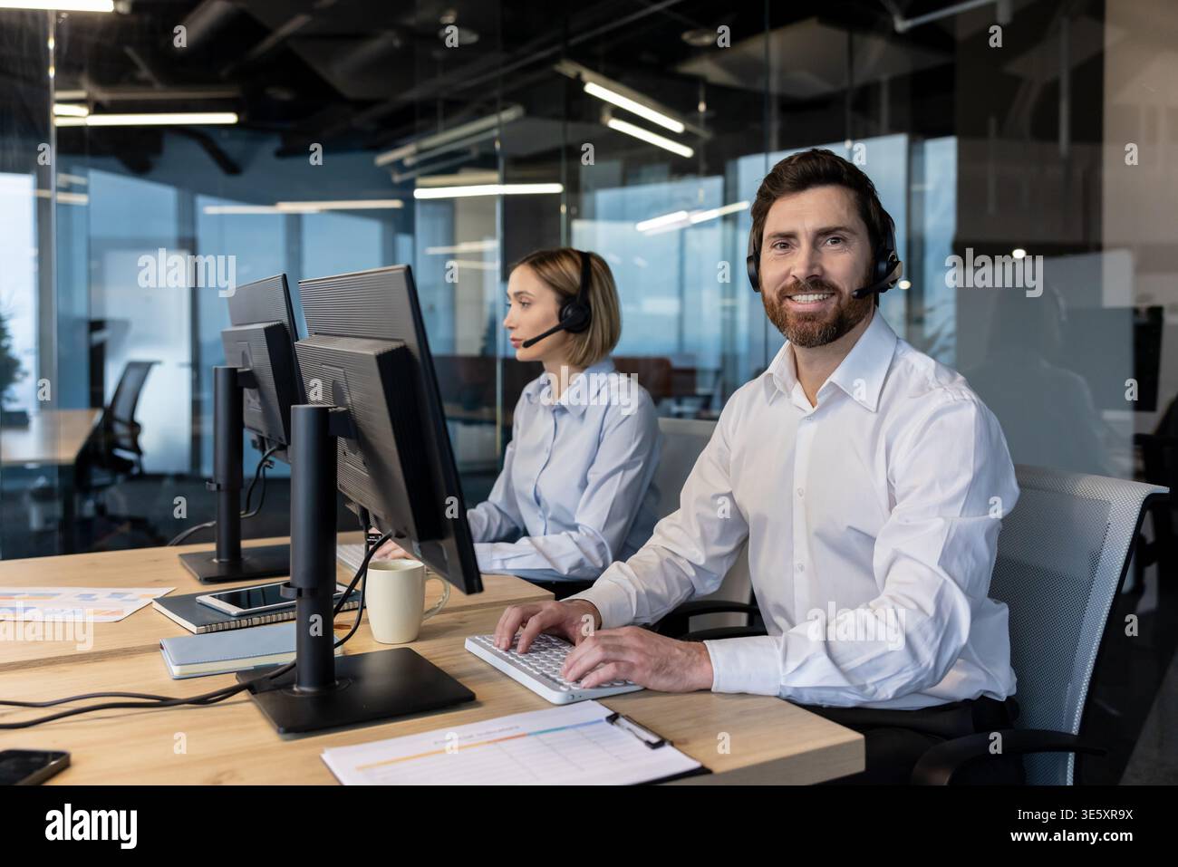 Un operatore sorridente di contact centre in un ufficio moderno che indossa una cuffia e lavora al computer, fornendo assistenza clienti mentre un collega lavora in background, lavoro di squadra professionale Foto Stock
