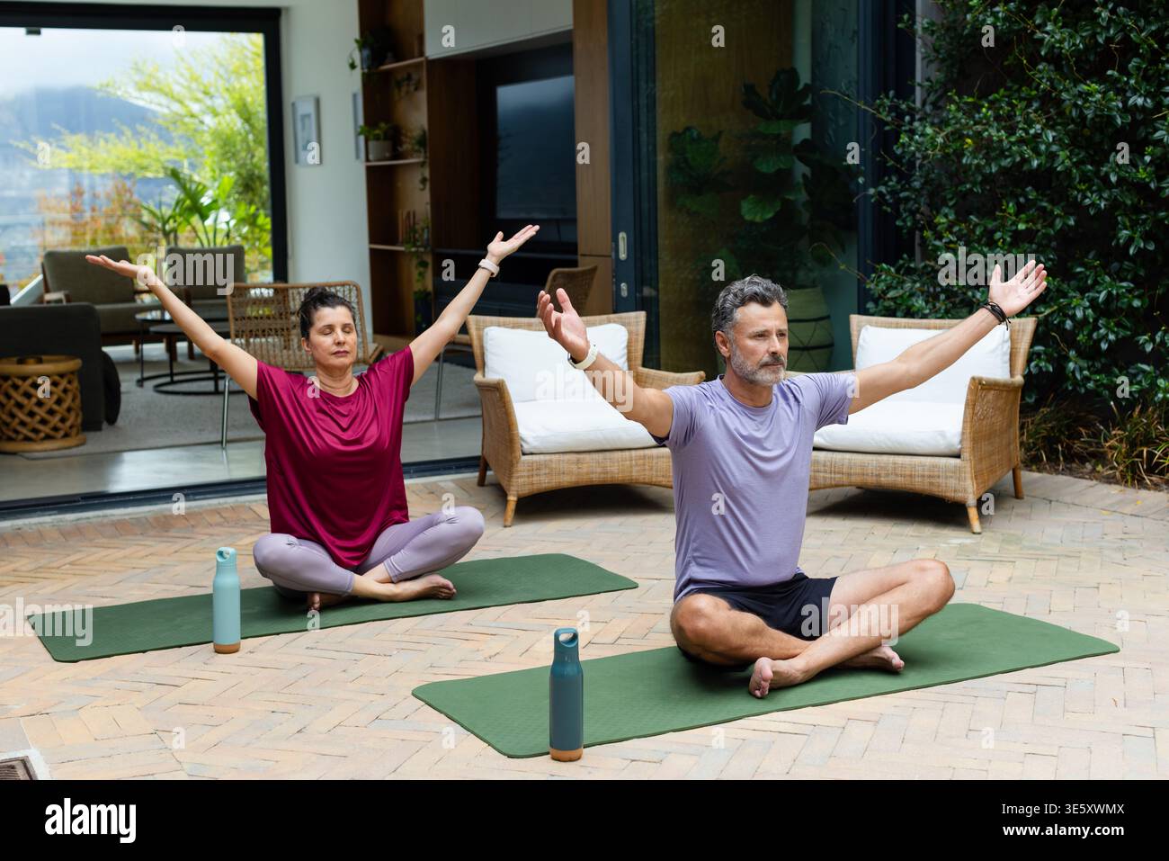 Coppie senior che praticano yoga su tappeti verdi nel patio in marrone e bottiglie viola chiaro e blu nelle vicinanze Foto Stock