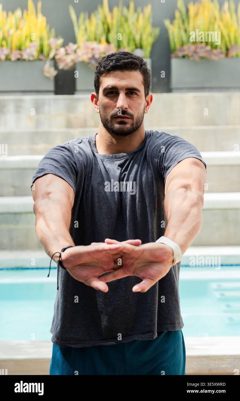Uomo adulto in piedi a bordo piscina sui gradini, indossa T-shirt scura, pantaloncini blu, fascia bianca, fioriere visibili Foto Stock