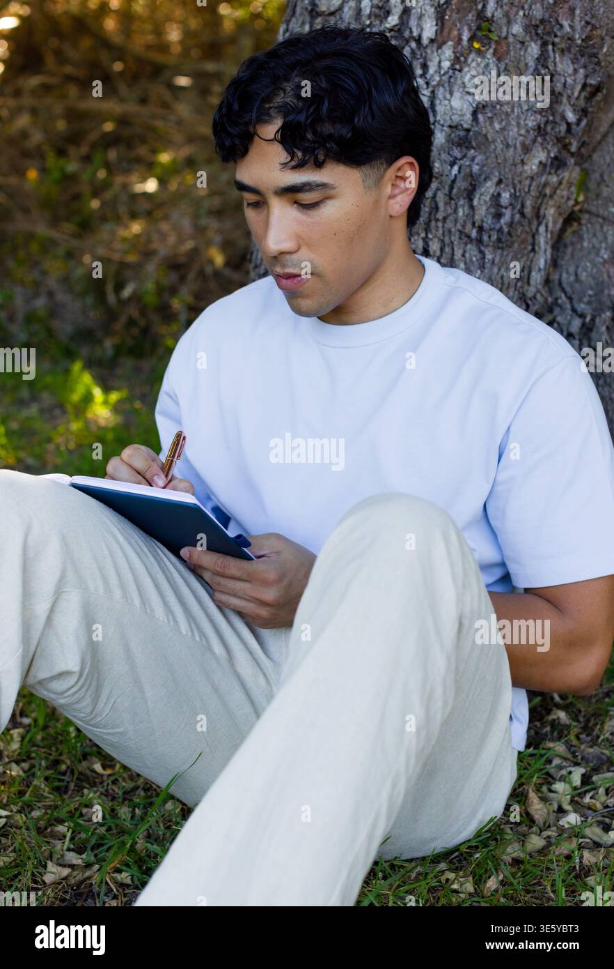 Uomo asiatico seduto sull'erba scrivendo in un taccuino con penna di metallo che indossa camicia azzurra, pantaloni beige Foto Stock