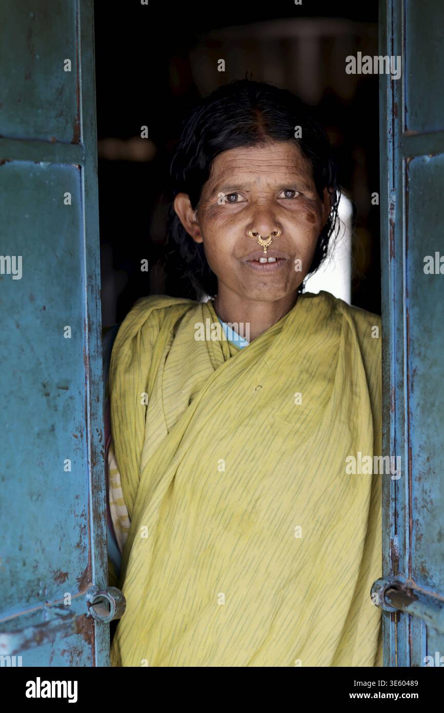 Odisha, India - 26 marzo 2026: Veduta di una donna locale incorniciata dalla porta intemprata e color verde acqua, la sua espressione serena un faro di resilienza e q Foto Stock