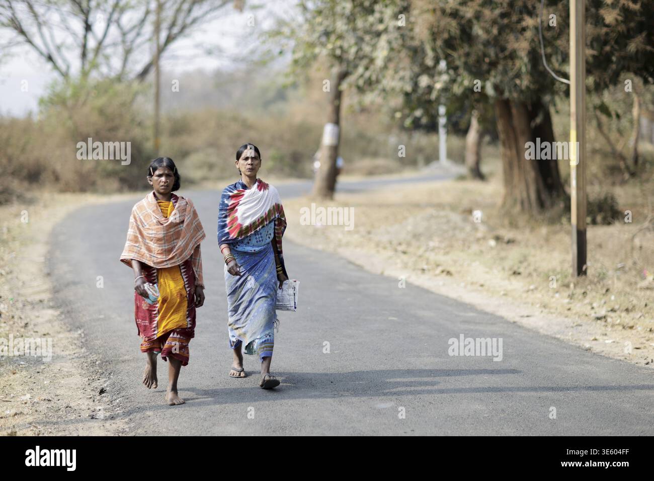 Odisha, India - 26 marzo 2026: Vista di due donne che camminano lungo una strada soleggiata, i loro saris colorati contrastano con i toni asciutti e silenziosi del Foto Stock