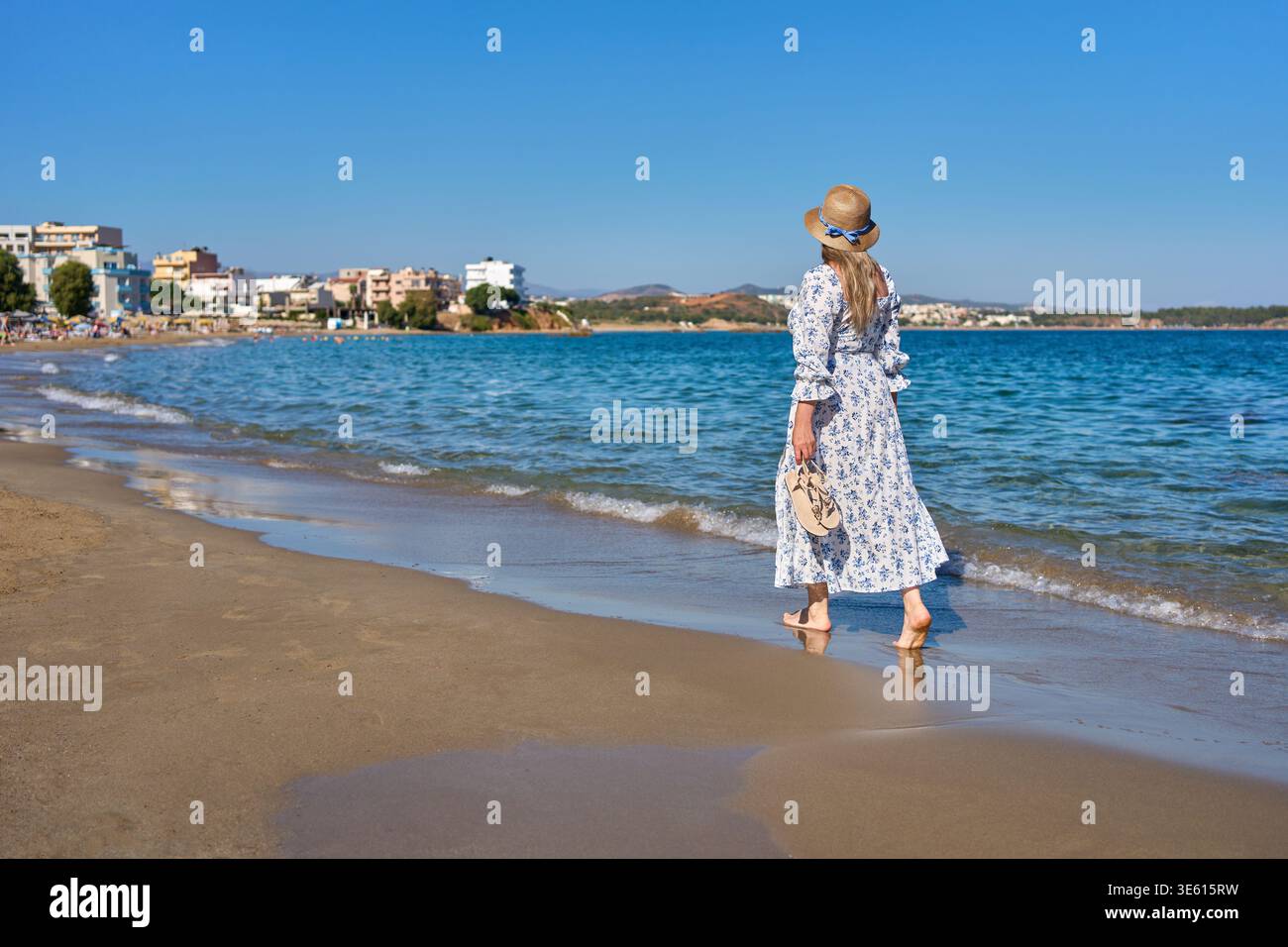Donna con maxi abito floreale e cappello di paglia che cammina a piedi nudi lungo una spiaggia sabbiosa a Chania, Creta. Splendido mare blu e sfondo della costa della città. Foto Stock