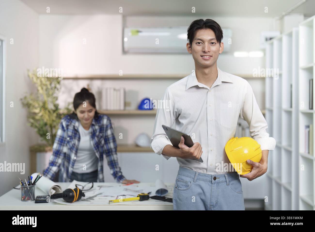 Un giovane ingegnere tiene in mano un tablet e un elmetto mentre discute i piani di progetto con un collega in uno spazio di lavoro moderno Foto Stock