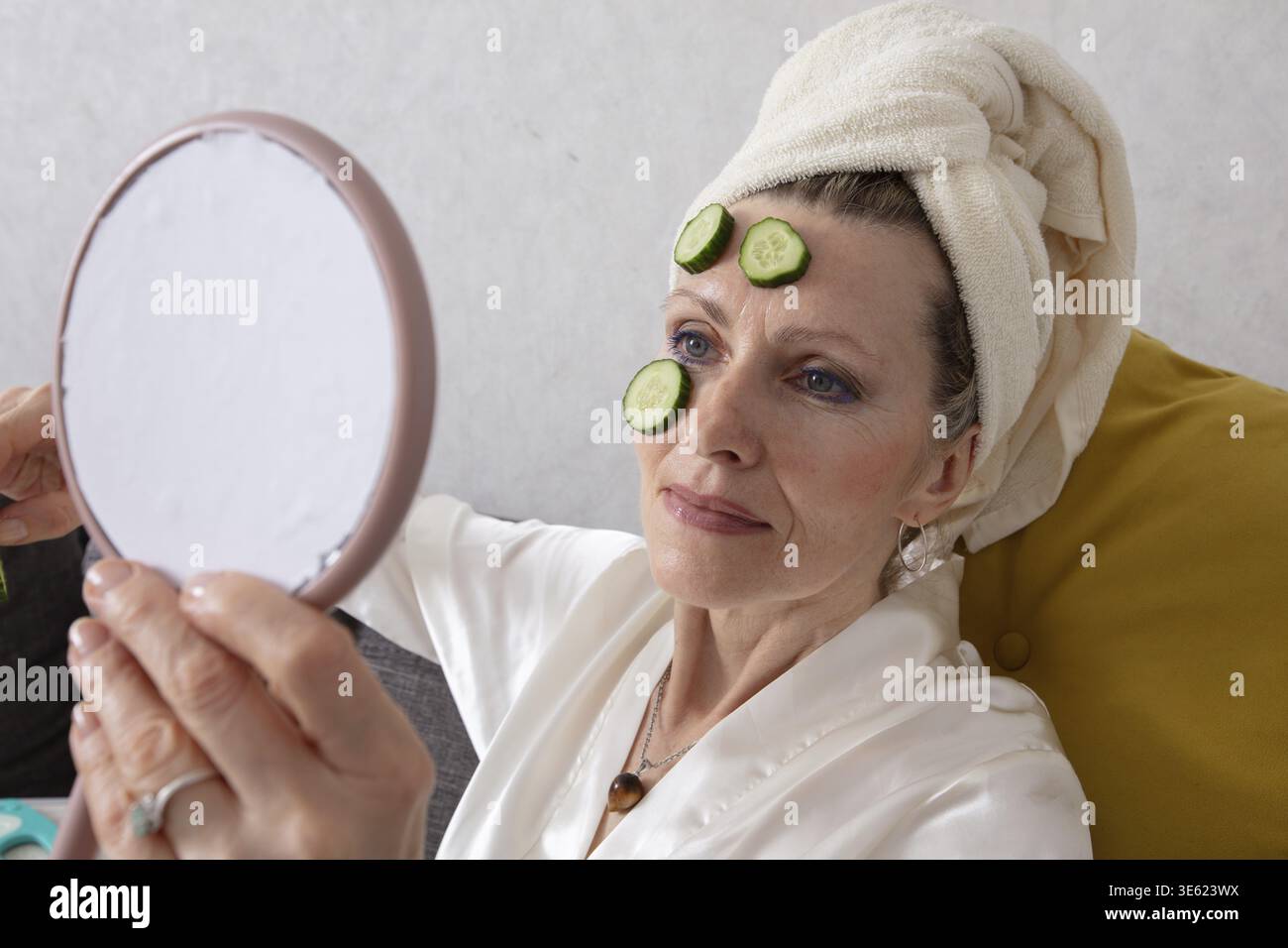 La donna anziana si siede comodamente sul divano avvolto in un asciugamano, applicando la cura della pelle con l'aiuto di fette di cetriolo, appoggiandosi sulla fronte, l Foto Stock