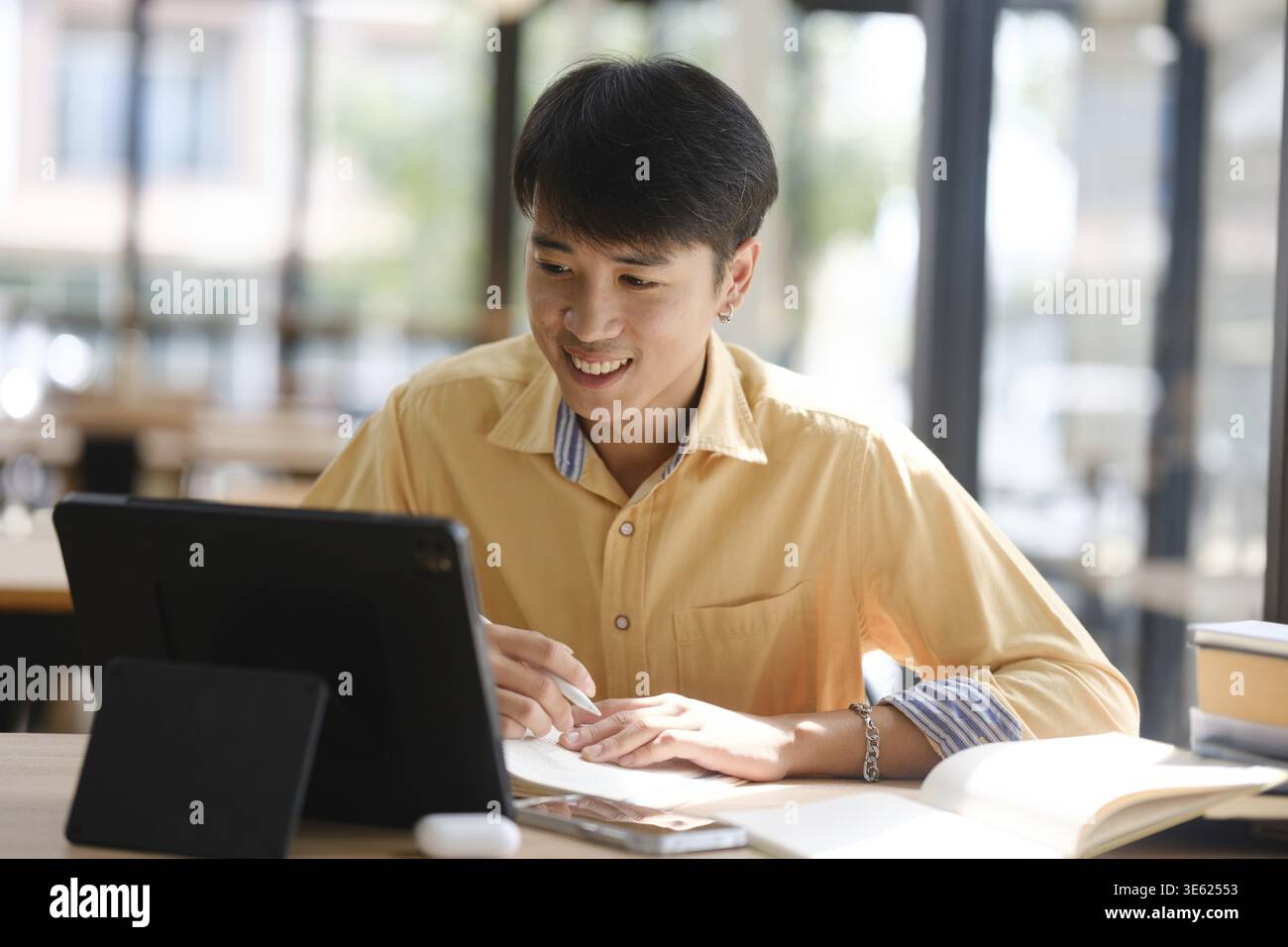 Uno studente impegnato nell'apprendimento online, prendendo appunti e rivedendo i materiali dello studio con libri e dispositivi digitali per prepararsi agli esami Foto Stock