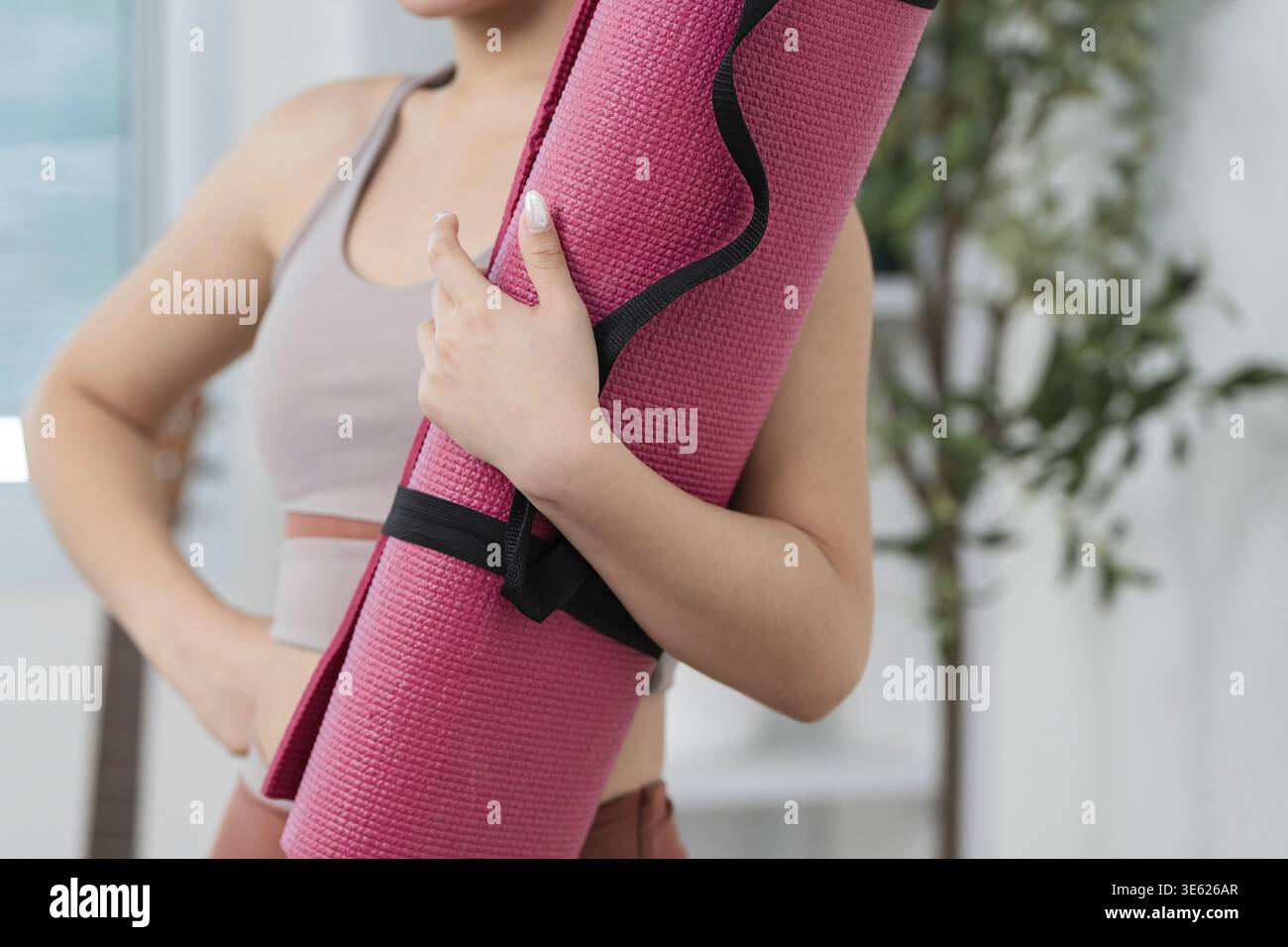 Una giovane donna sicura di sé tiene il suo tappetino da yoga rosa, preparandosi per l'allenamento in un ambiente domestico sereno Foto Stock