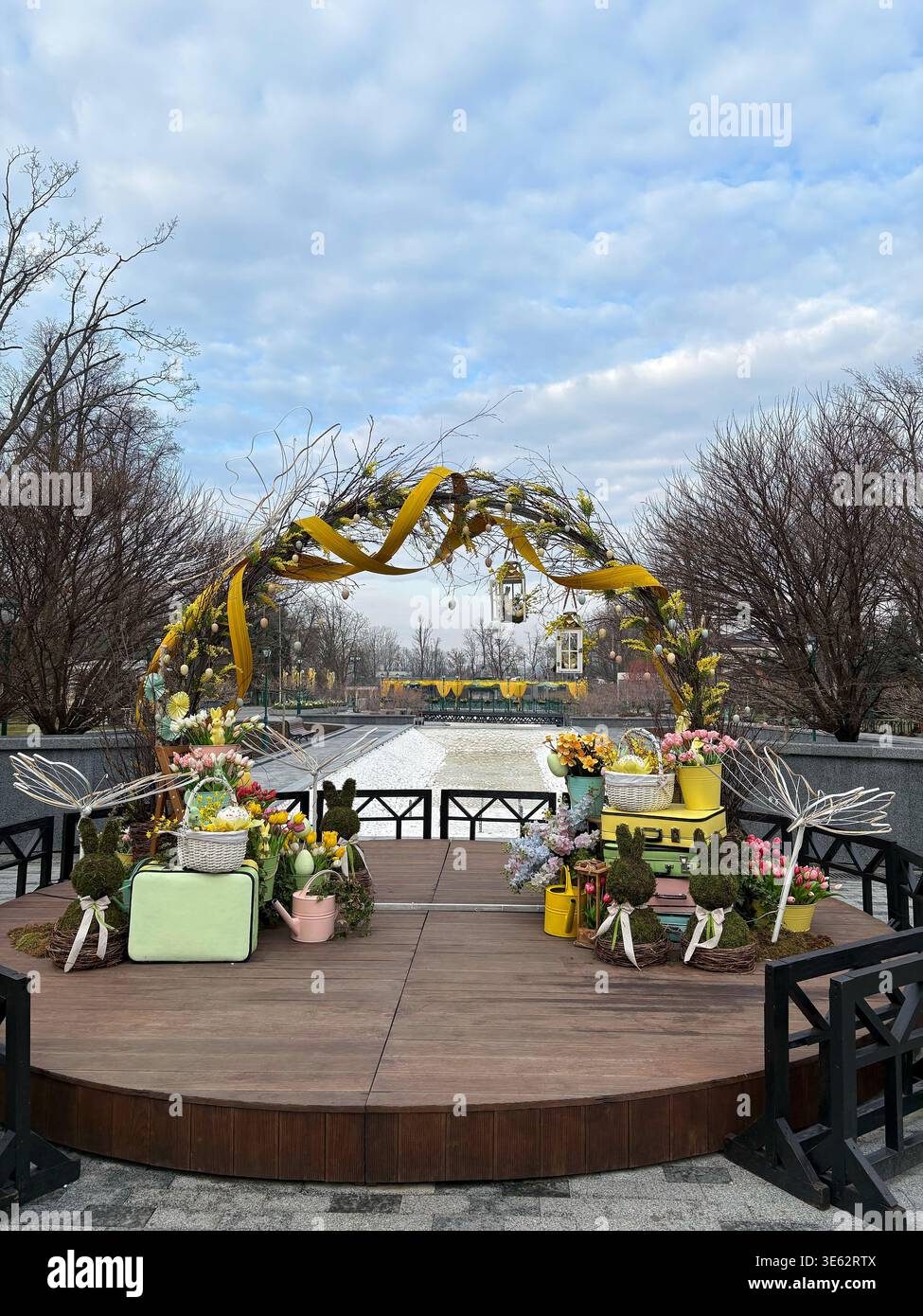 Una scenografica mostra pasquale all'aperto adagiata su una piattaforma di legno in un parco pubblico, caratterizzata da un arco decorato con nastri gialli, composizioni floreali e muschio Foto Stock