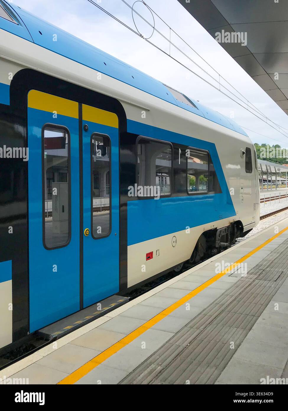 Frammento di un treno passeggeri di un operatore ferroviario slovacco in una stazione con cavi aerei, linea di sicurezza gialla e piattaforma vuota Foto Stock