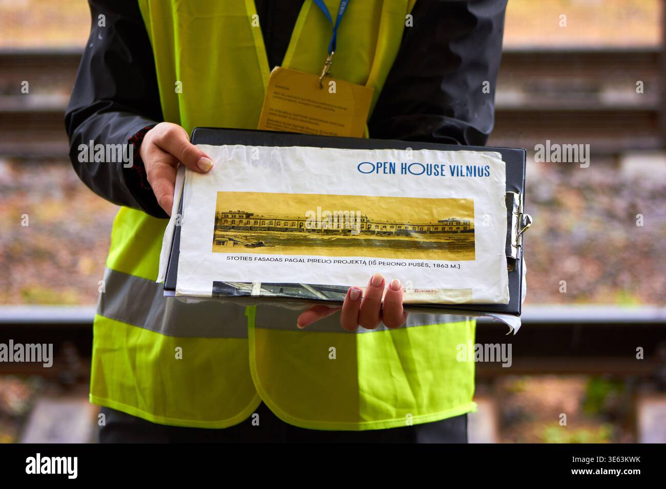 Una persona che indossa un giubbotto di sicurezza giallo ad alta visibilità e una giacca nera tiene in mano un portadocumenti che mostra la documentazione architettonica. Il documento in alto Foto Stock
