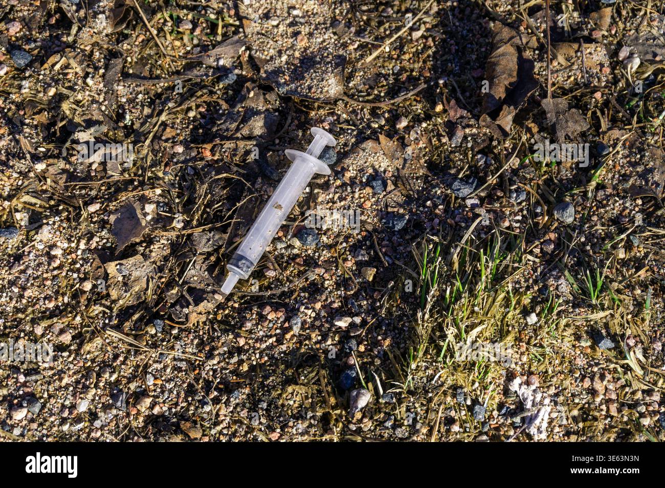 Una siringa usata giace su un terreno sabbioso e grintoso tra piccole rocce e erba sparsa, evidenziando l'inquinamento urbano. Foto Stock
