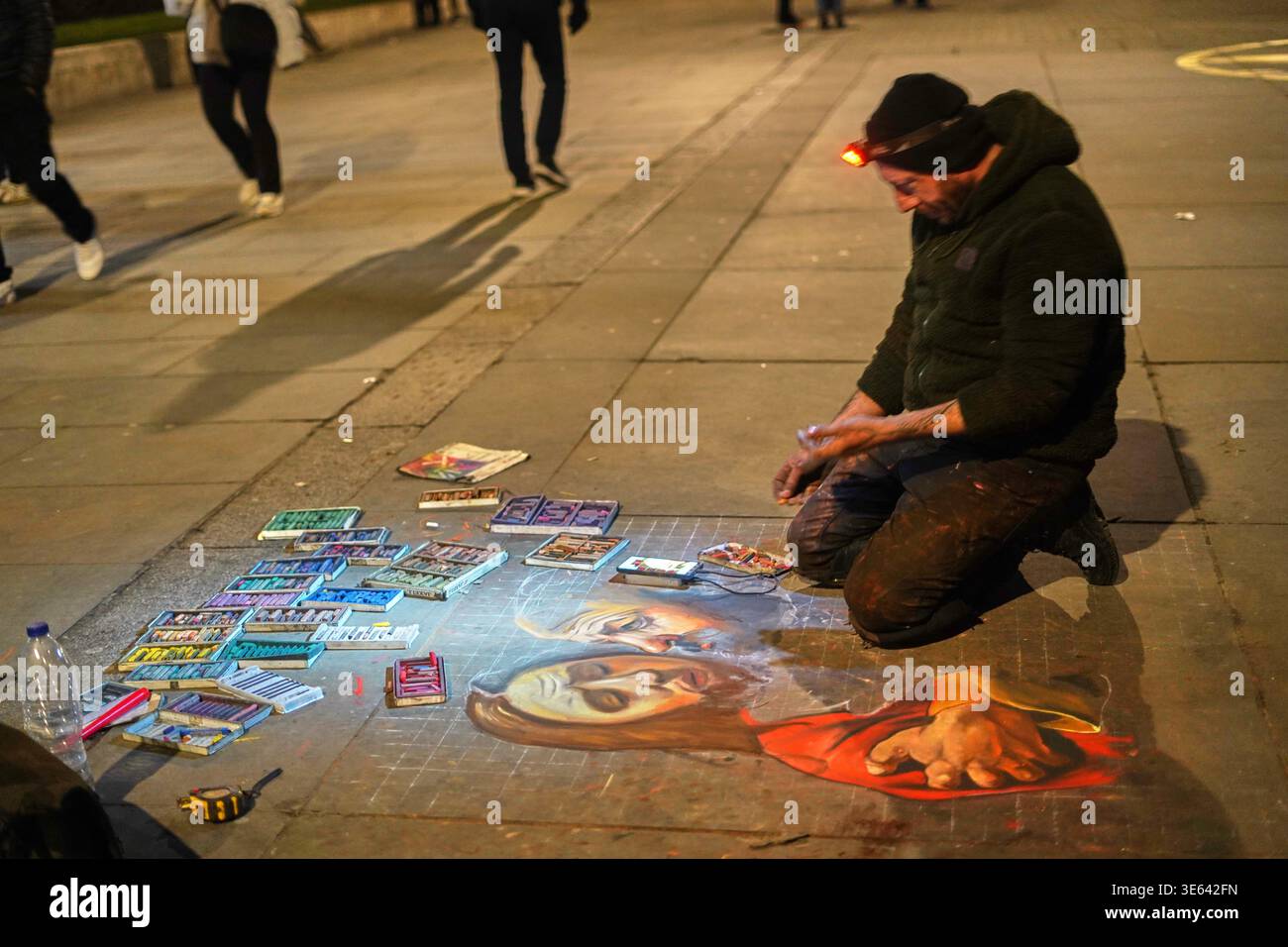 Un artista di strada si inginocchia su un marciapiede pavimentato di notte creando un grande disegno in gesso. L'artista indossa un berretto nero con un arancione illuminato Foto Stock