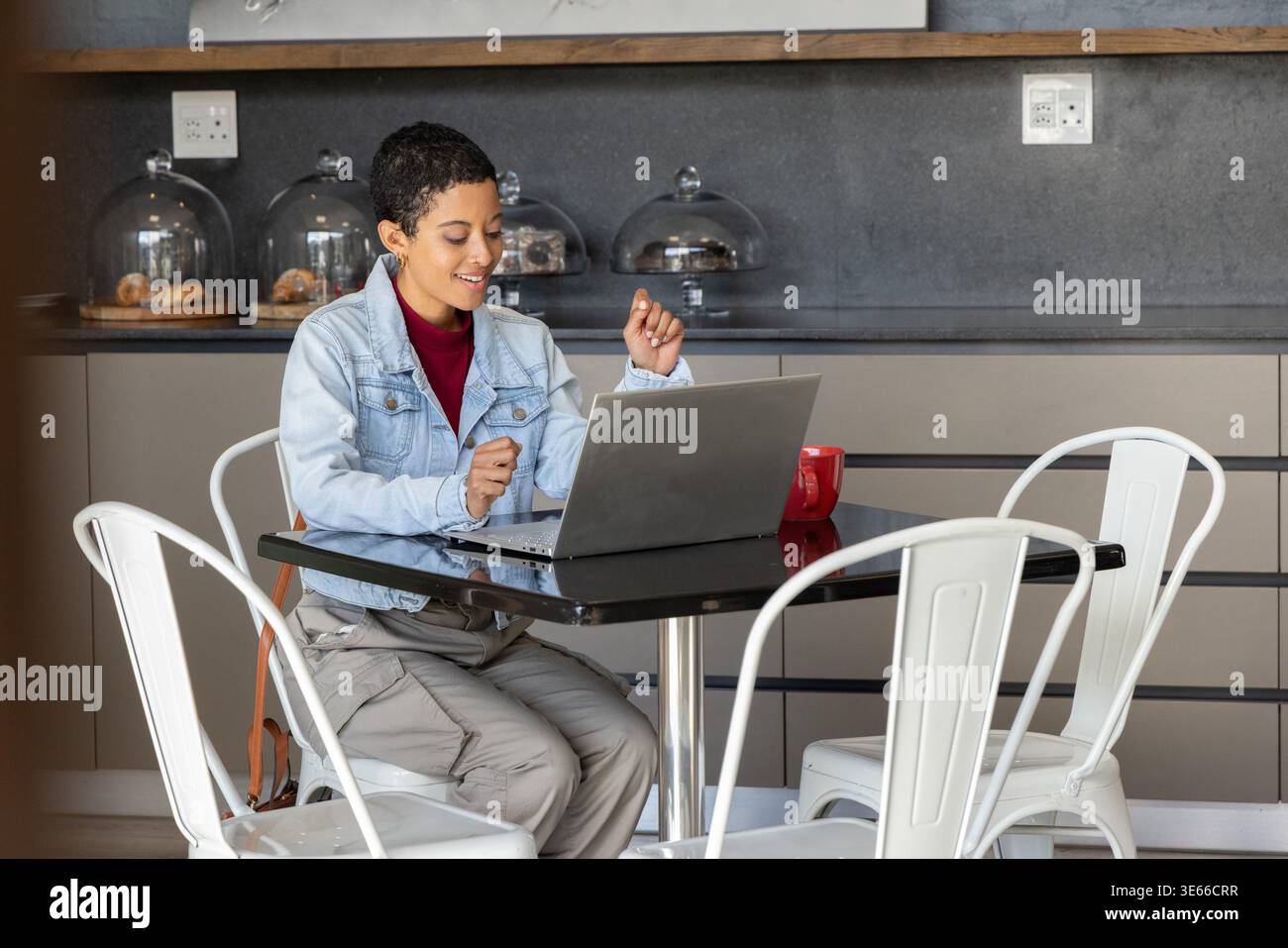 Donna afroamericana con camicia in denim seduta al bar che lavora su un computer portatile con tazza rossa e pasticcini Foto Stock