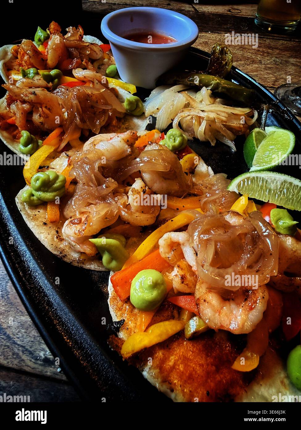 Tacos di gamberi freschi serviti su un piatto con peperoni grigliati, cipolle, crema di avocado, spicchi di lime e salsa su un tavolo di legno Foto Stock