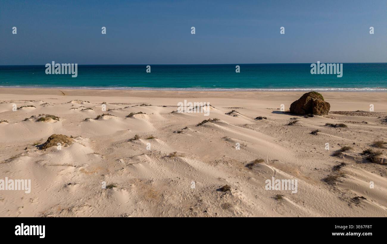 Spiaggia di sabbia vuota con dune scolpite dal vento, orizzonte turchese del mare e cielo azzurro cristallino, vista mare minima con rocce in primo piano, spazio per i viaggi Foto Stock