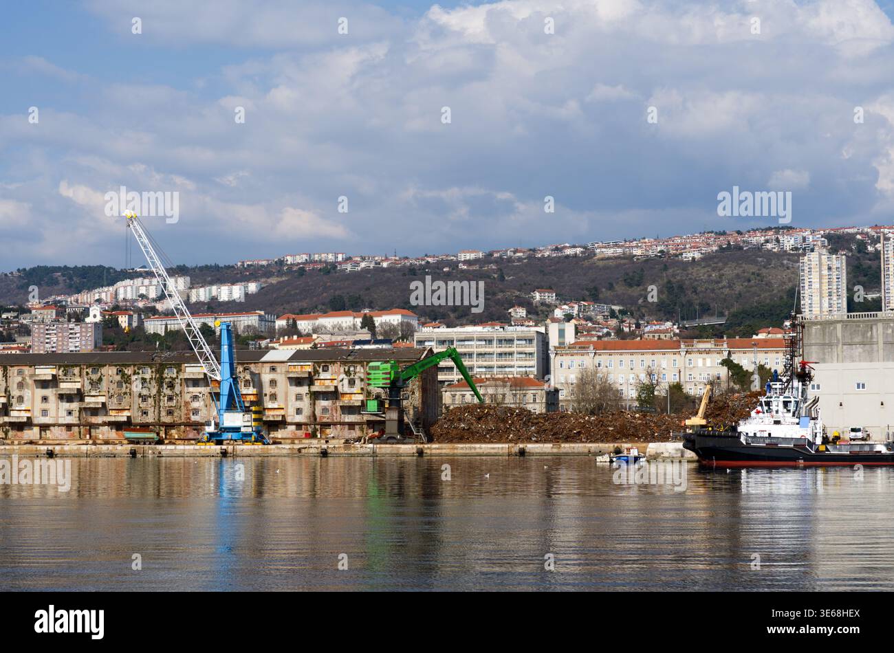 Vista di un porto industriale commerciale con gru da carico blu e verdi per impieghi gravosi, un grande cumulo di riciclaggio dei rottami metallici sul molo e il vecchio porto Foto Stock