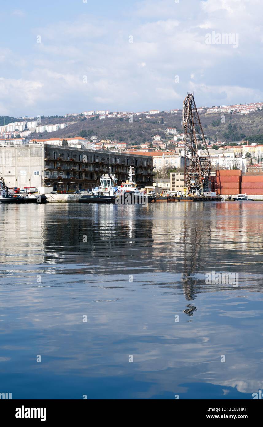 Una zona portuale caratterizzata da acque calme e riflettenti, barche da lavoro attraccate e una grande gru industriale vicino al lungomare. Si erge un edificio portuale in cemento Foto Stock