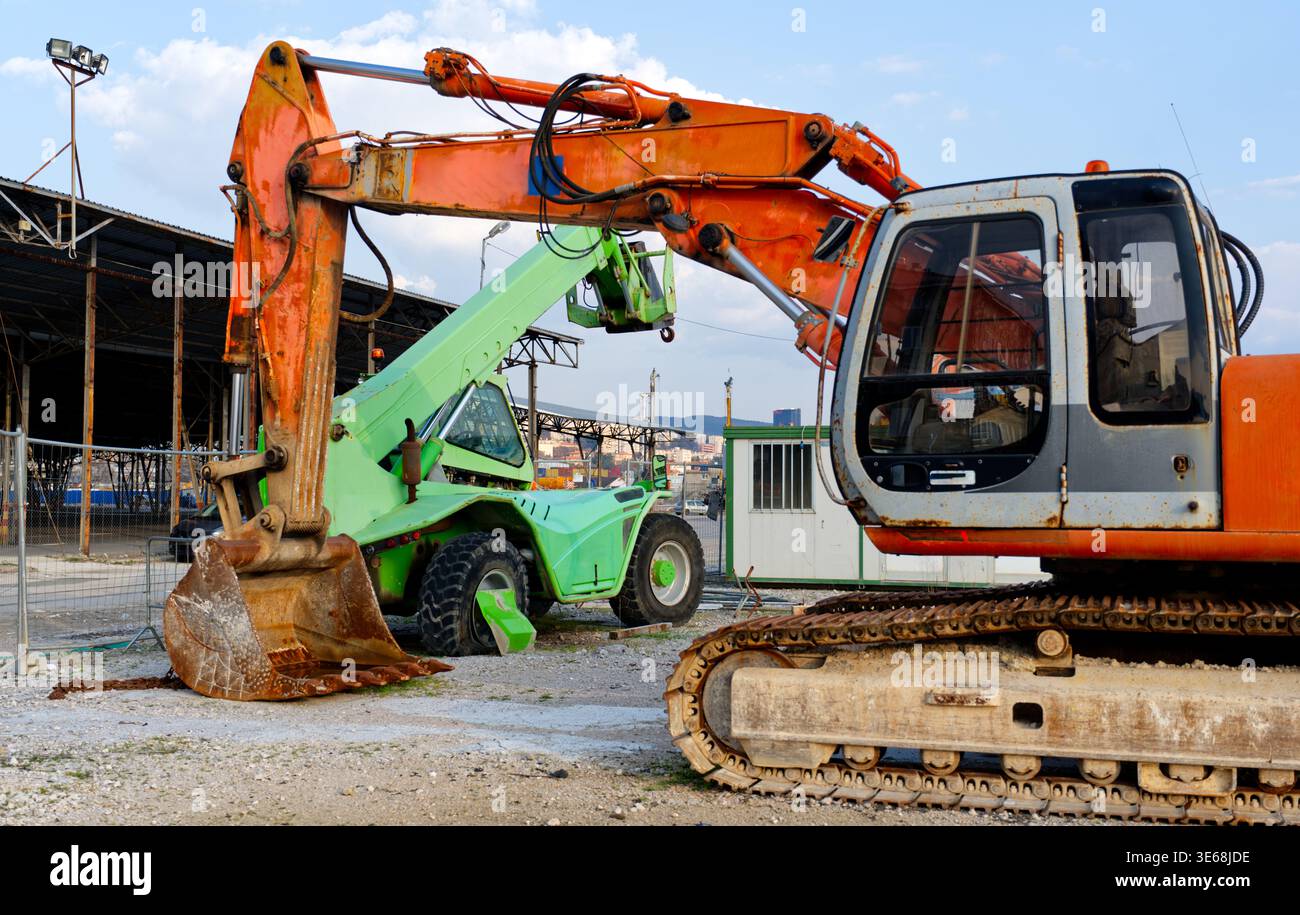 Escavatore cingolato arancione e movimentatore telescopico verde calce parcheggiato in un cantiere o in un cantiere industriale. Macchinari pesanti e attrezzature di movimento terra Foto Stock