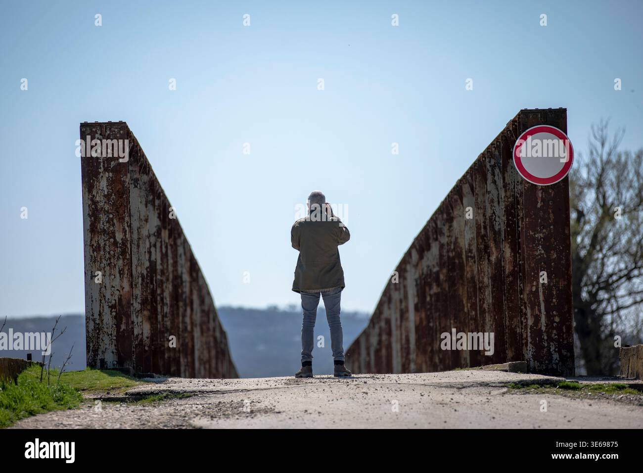 Uomo in piedi su un vecchio ponte arrugginito, a fare una foto. La forte simmetria e le linee principali creano una composizione minimalista sotto un cielo azzurro. Foto Stock