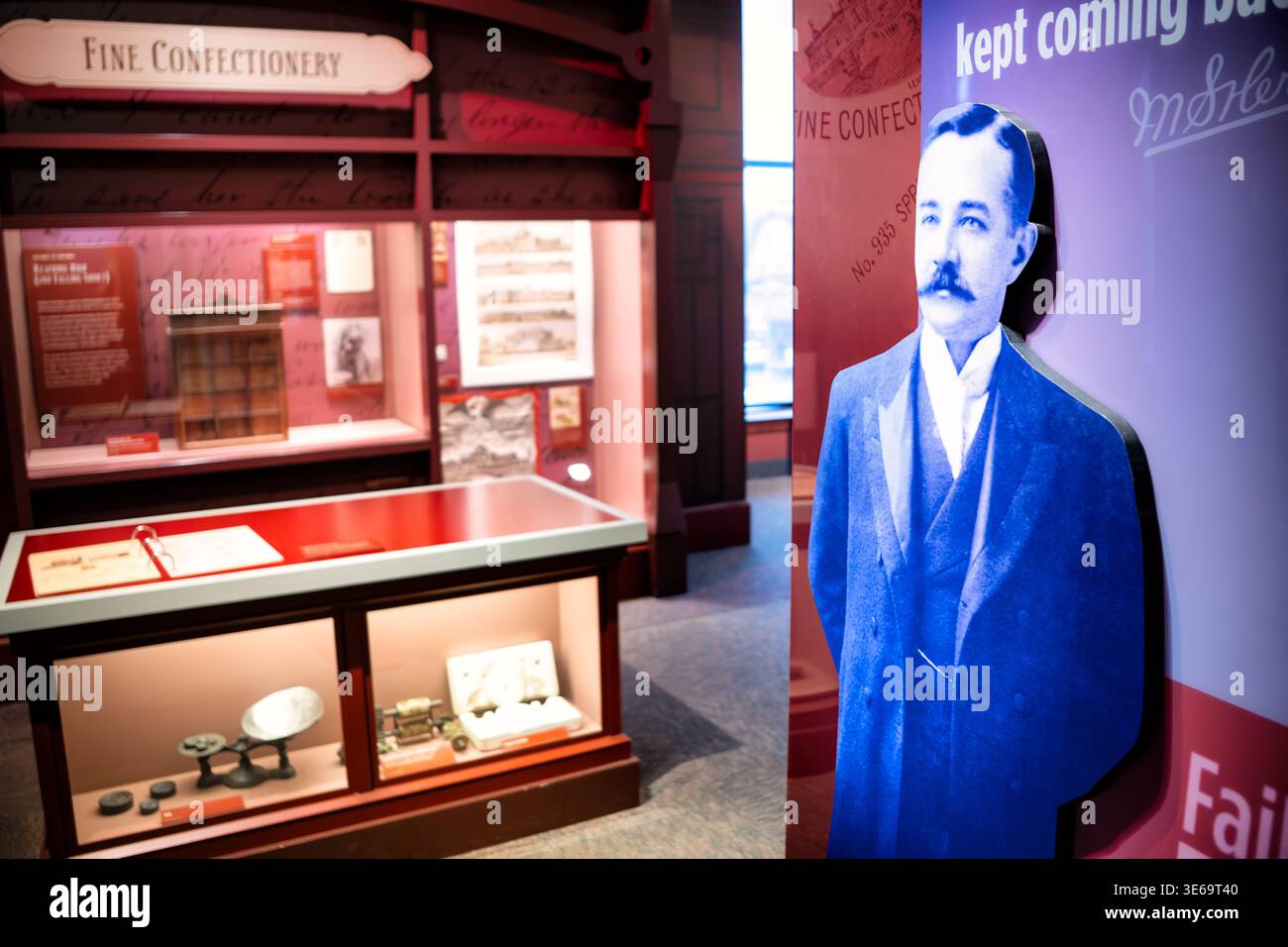 Hershey Story Museum Esposizione del ritratto di Milton Hershey Hershey Stati Uniti | HERSHEY, Pennsylvania — Un ritratto monocromatico ritagliato di Milton Hershey si trova accanto a vetrine illuminate con artefatti antichi della produzione di dolciumi all'interno del The Hershey Story Museum. L'esposizione racconta il percorso dell'industriale, dalle sue prime imprese di confetteria senza successo alla fondazione del suo impero mondiale del cioccolato. Artefatti curati, tra cui bilance meccaniche d'epoca e stampi per dolci, illustrano i processi di fabbricazione della fine del XIX secolo che hanno preceduto la produzione di massa dell'iconico Foto Stock