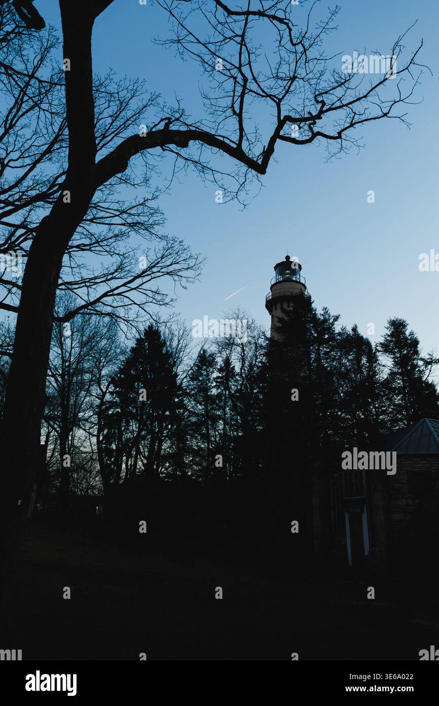 Un faro è visibile al crepuscolo contro un cielo blu. Alberi alti circondano il faro mentre i loro rami si allungano verso il cielo. Un debole aereo tra Foto Stock
