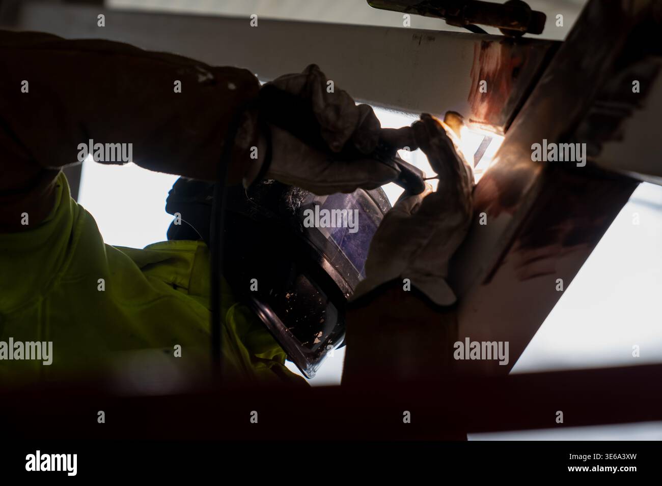 Lavori di saldatura industriale sotto un telaio in acciaio con visiera protettiva e guanti, evidenziando la costruzione di metalli, l'assemblaggio e la manodopera specializzata Foto Stock