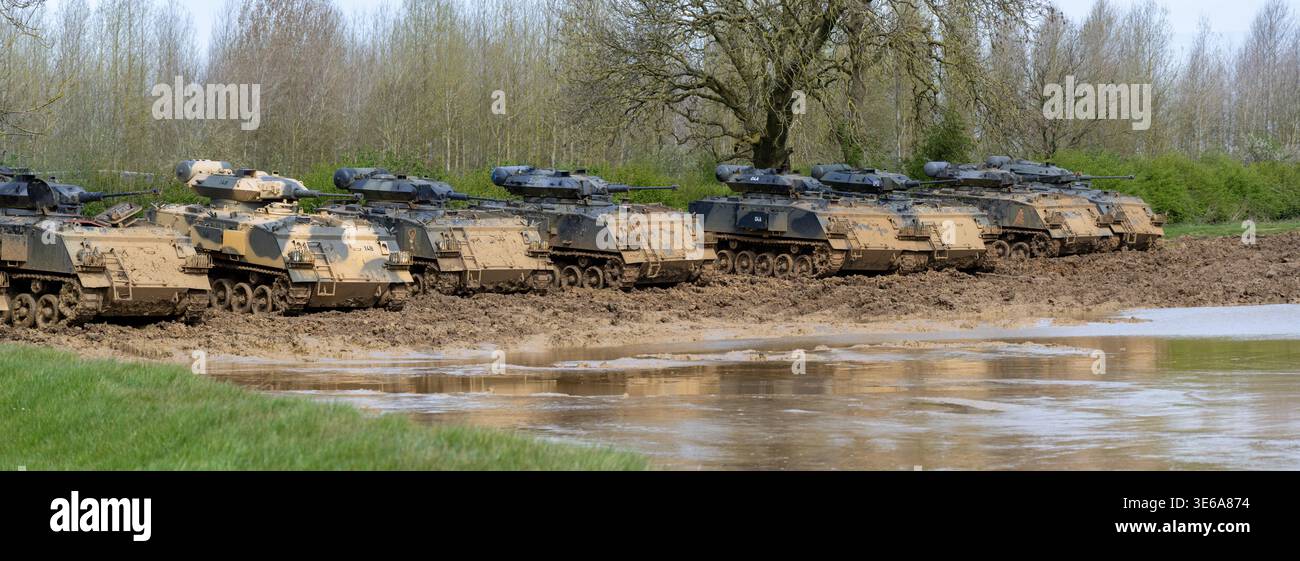 Vista panoramica di più veicoli blindati allineati su un terreno di addestramento fangoso accanto all'acqua, che illustra le moderne forze meccanizzate a Armourgeddon mi Foto Stock