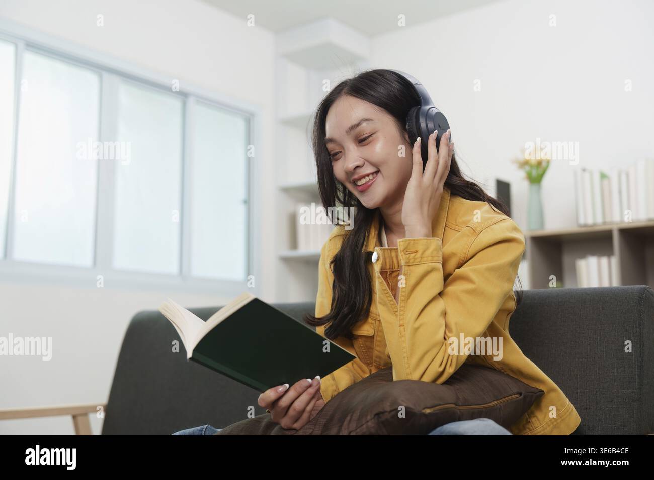 Una donna con le cuffie legge un libro, godendosi un momento tranquillo di relax e cura di sé nella sua accogliente casa Foto Stock