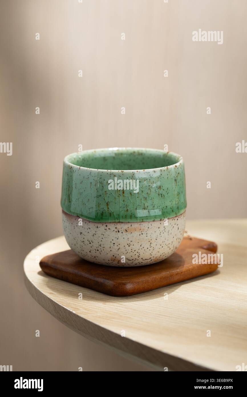 Tazza da tè in ceramica giapponese minimalista collocata su un sottobicchiere in legno con luce naturale soffusa, a creare un'atmosfera tranquilla e intima su un tavolo in legno. Foto Stock