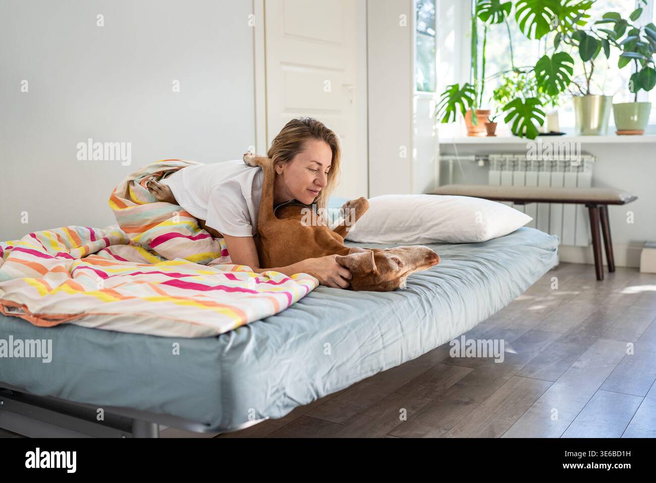 Donna positiva che si coccola con l'amato cane Vizsla in un comodo letto di casa, mostrando amore e armonia. Foto Stock
