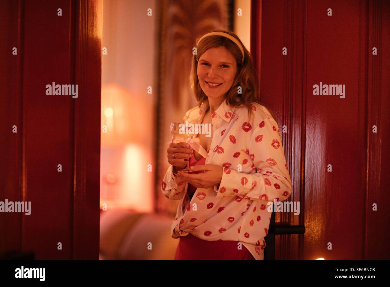Una giovane donna con una camicia a motivi geometrici si erge su un portone che regge un vetro, con un interno delicatamente illuminato caratterizzato da colori caldi ed elementi decorativi nella Foto Stock