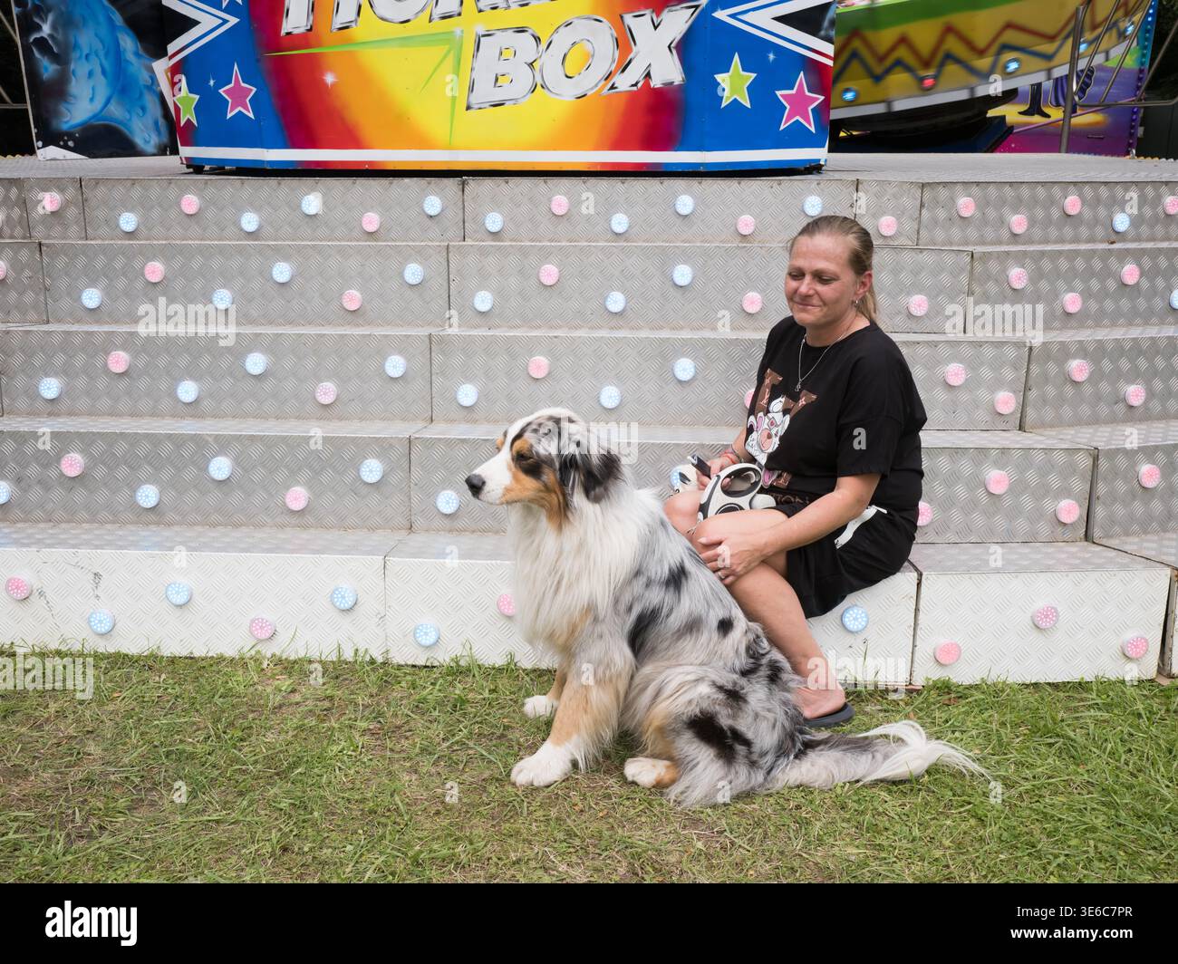 Donna di mezza età seduta con un cane da pastore australiano di fronte a un'attrazione per il divertimento durante il giorno Foto Stock