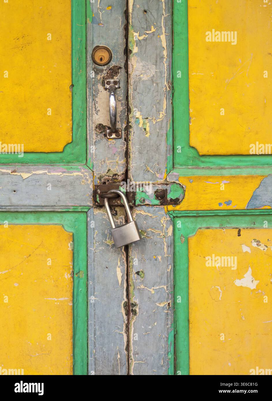 Primo piano di una vecchia porta in legno gialla con vernice sbucciata, maniglia in metallo, serratura e lucchetto argento su finiture verde-blu Foto Stock