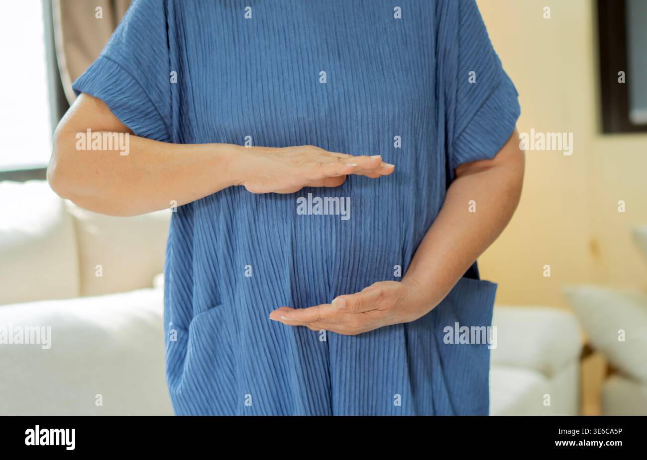 Foto di una donna anziana non identificabile che indossa un vestito blu che posa con le mani sullo stomaco nel salotto. Concetto di stomaco. Copia spazio. Foto Stock