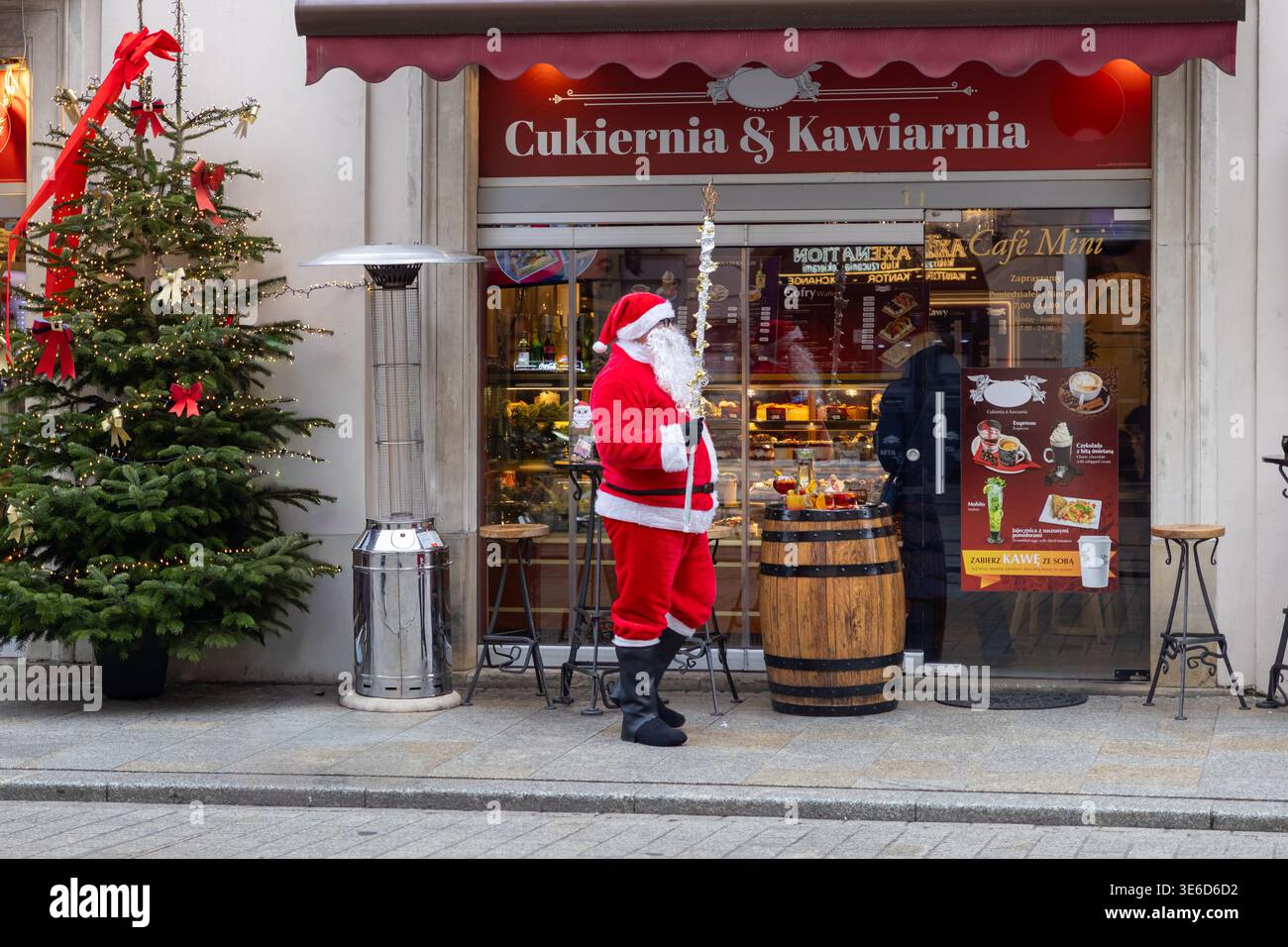 Cracovia, Polonia - 2 dicembre 2025: Babbo Natale si erge sul marciapiede della città, accanto a un albero di Natale decorato e a un vivace caffè, che irradia una calda vacanza festiva Foto Stock