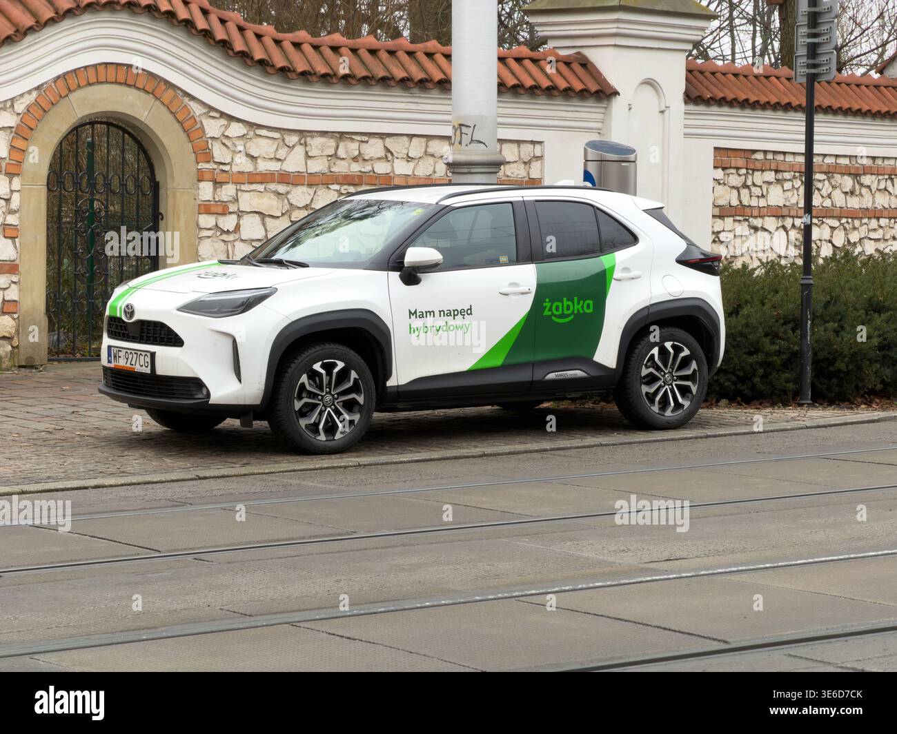 Cracovia, Polonia - 2 dicembre 2025: Un'auto ibrida Zabka bianca è parcheggiata in una strada acciottolata accanto ai binari del tram, promuovendo un trasporto urbano ecologico Foto Stock