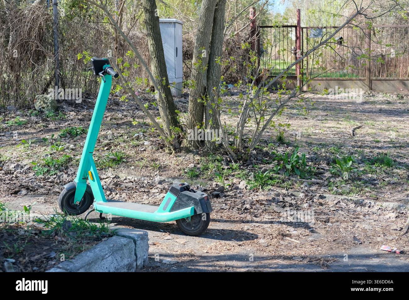 Lo scooter elettrico è parcheggiato su un sentiero sterrato vicino ad alberi e cancelli metallici in un'area residenziale all'aperto Foto Stock