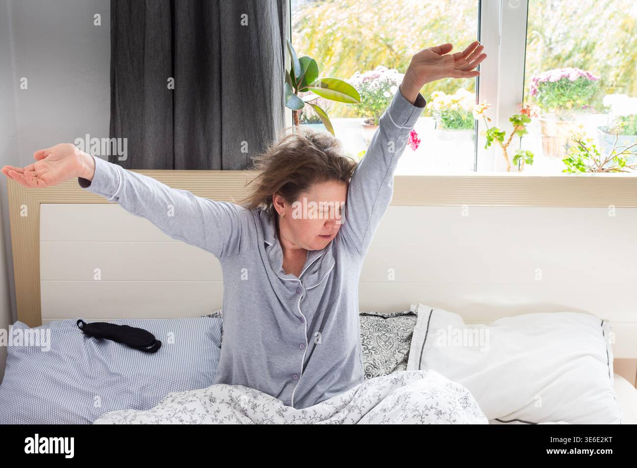 Una donna di mezza età si siede a letto, allungando le braccia sopra la testa. Sembra stanca e non ha dormito abbastanza. La sala è luminosa e naturale Foto Stock