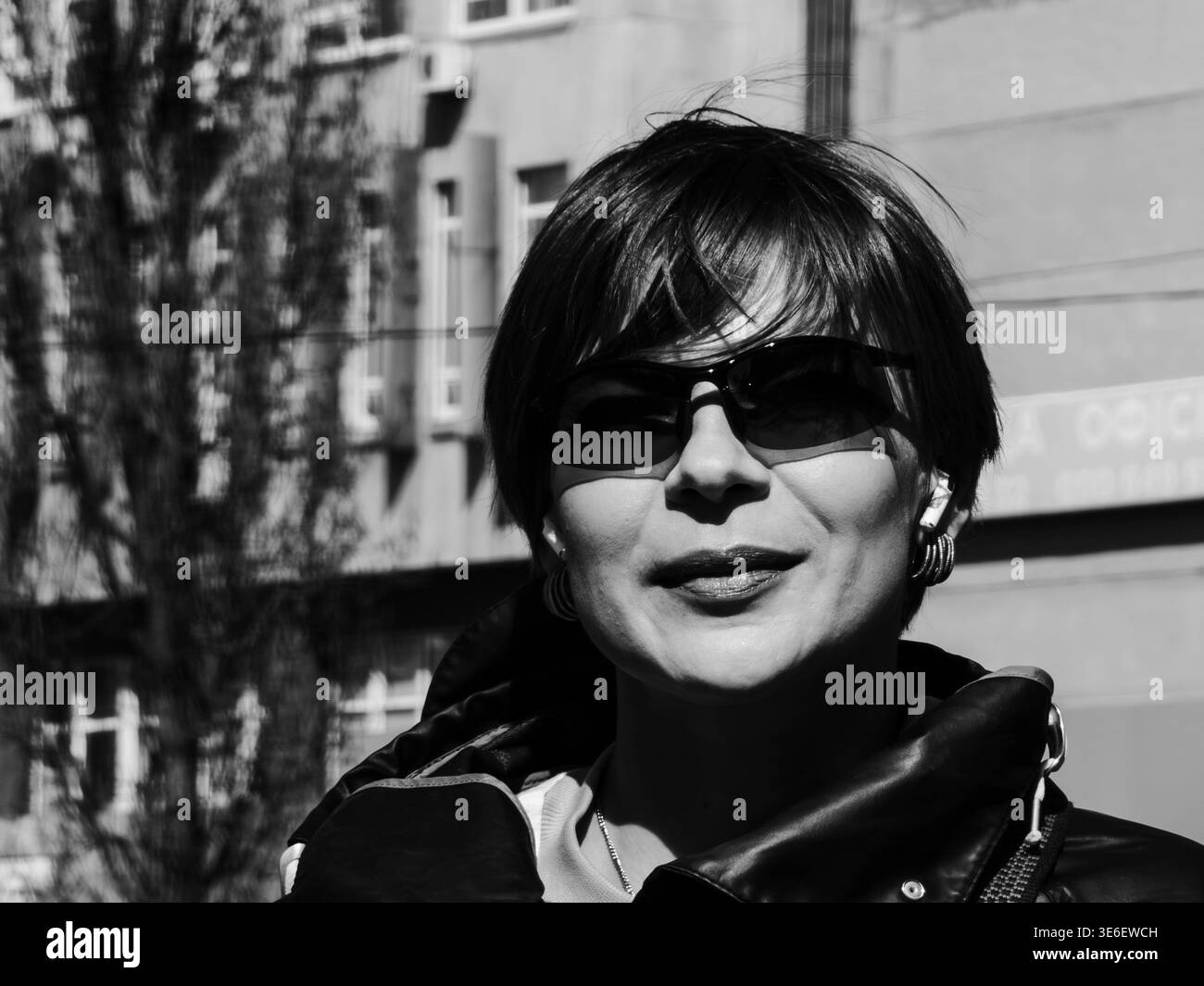 Donna che indossa occhiali da sole e un'espressione sicura mentre cammina all'aperto su una strada urbana, il suo viso è parzialmente ombreggiato dalla luce del sole e crea un forte cont Foto Stock