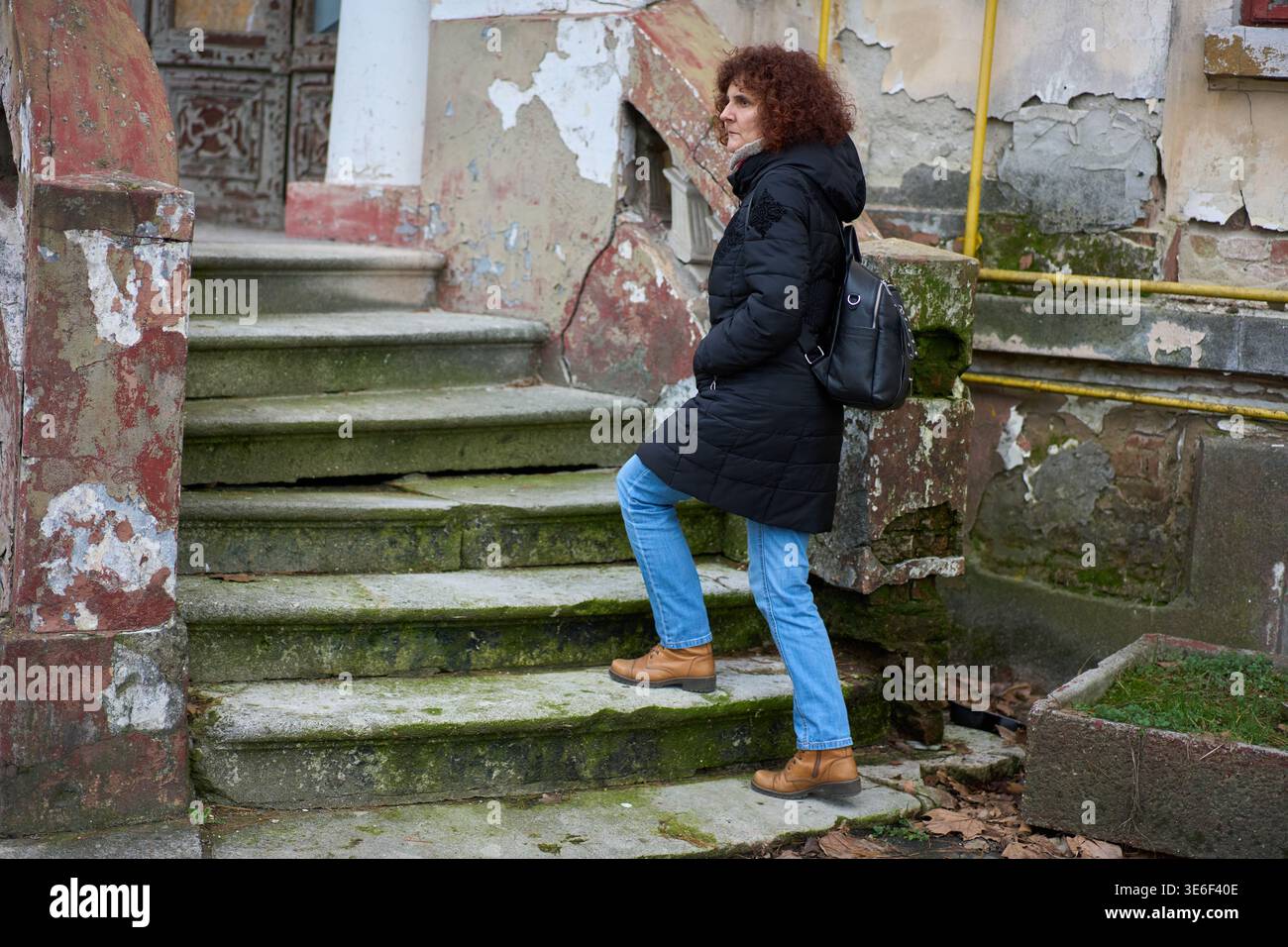 Donna matura con capelli rossi ricci che salgono scalini in pietra ricoperti di muschio di un edificio storico in decadenza con pareti dipinte in pelatura in un giorno d'inverno Foto Stock